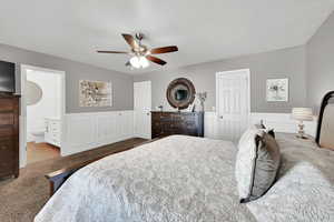 Bedroom with a ceiling fan, carpet flooring, a wainscoted wall, a textured ceiling, and connected bathroom