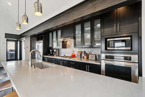 Kitchen featuring appliances with stainless steel finishes, dark cabinets, decorative backsplash, decorative light fixtures, and glass insert cabinets