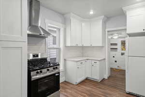 Kitchen featuring stainless steel range with gas cooktop, freestanding refrigerator, wall chimney exhaust hood, dark wood-style flooring, and white cabinets