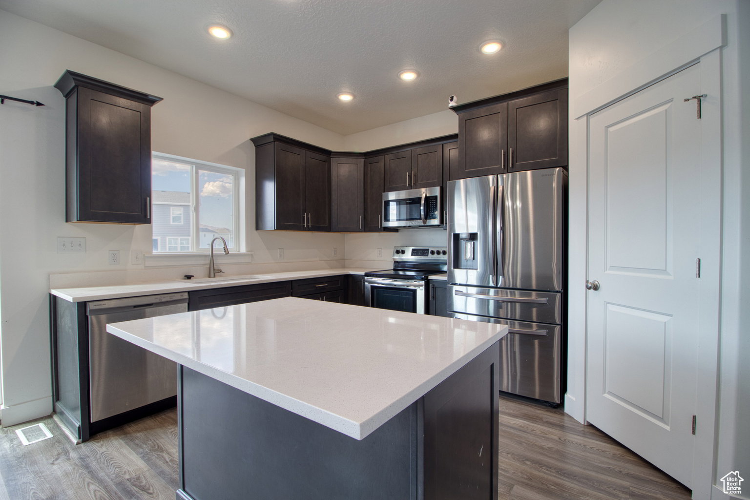 Kitchen with appliances with stainless steel finishes, dark brown cabinetry, dark wood-style flooring, light stone counters, and recessed lighting
