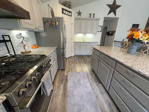 Kitchen featuring gray cabinetry, stainless steel stove, under cabinet range hood, light stone counters, and lofted ceiling