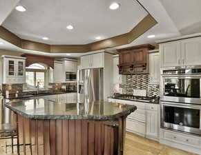 Kitchen featuring a trey ceiling, stainless steel appliances, a kitchen breakfast bar, glass cabinets, and dual colored custom cabinets.
