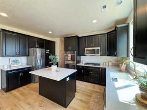 Kitchen with light wood-style floors, appliances with stainless steel finishes, dark cabinetry, a textured ceiling, and recessed lighting