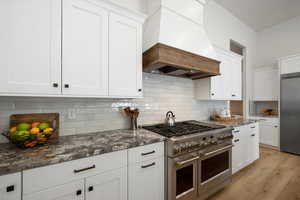 Kitchen with high end appliances, backsplash, dark stone counters, white cabinetry, and custom range hood