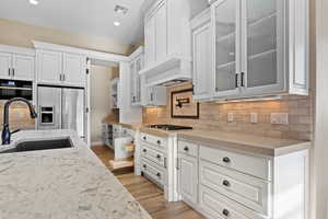 Kitchen with white cabinets, decorative backsplash, light stone counters, glass insert cabinets, and recessed lighting