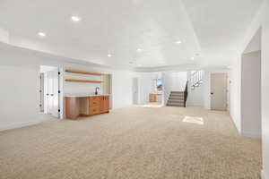 Unfurnished living room featuring light colored carpet, recessed lighting, stairs, bar with sink, and a tray ceiling