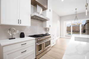 Kitchen with double oven range, white cabinetry, wall chimney exhaust hood, light stone counters, and decorative light fixtures