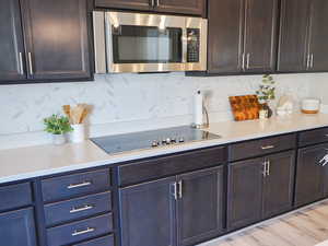 Kitchen featuring decorative backsplash, dark brown cabinets, stainless steel microwave, black electric stovetop, and light stone counters
