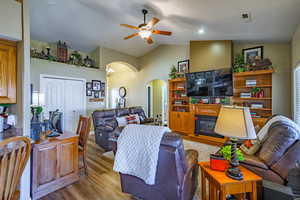 Living room with vaulted ceiling, light wood-style floors, arched walkways, a glass covered fireplace, and a ceiling fan
