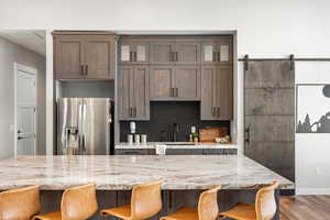 Kitchen featuring light stone countertops, tasteful backsplash, stainless steel refrigerator with ice dispenser, dark brown cabinets, and a barn door