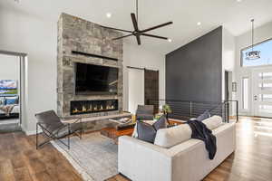 Living area with a barn door, a ceiling fan, a towering ceiling, wood-type flooring, and a stone fireplace