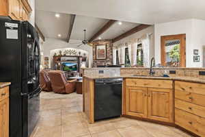 Kitchen with brown cabinetry, open floor plan, black appliances, light stone countertops, and beamed ceiling