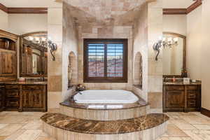 Full bathroom featuring stone tile flooring, two vanities, a garden tub, crown molding, and a chandelier