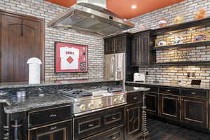 Kitchen featuring dark brown cabinets, dark stone countertops, ventilation hood, brick wall, and appliances with stainless steel finishes