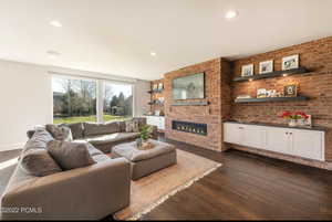 Living area with dark wood-type flooring, brick wall, a glass covered fireplace, and recessed lighting