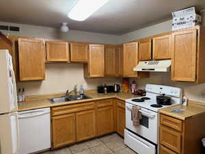 Kitchen featuring white appliances, light countertops, under cabinet range hood, light tile patterned flooring, and brown cabinetry