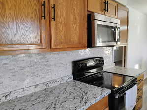 Kitchen with tasteful backsplash, black range with electric stovetop, brown cabinetry, stainless steel microwave, and light stone counters