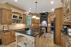 Kitchen with open shelves, decorative light fixtures, a center island with sink, brown cabinetry, and dark stone counters