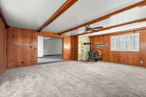 Unfurnished living room featuring a wood stove, wooden walls, beam ceiling, carpet flooring, and a ceiling fan