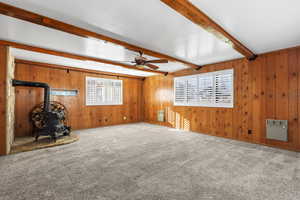 Carpeted empty room with a wood stove, wooden walls, beamed ceiling, heating unit, and ceiling fan