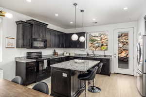 Kitchen with black appliances, light stone countertops, hanging light fixtures, a kitchen breakfast bar, and light wood finished floors