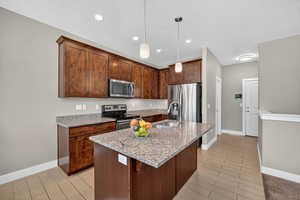 Kitchen with light stone counters, appliances with stainless steel finishes, an island with sink, hanging light fixtures, and wood finish floors