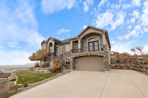 View of front of property with a balcony, stone siding, stucco siding, concrete driveway, and a garage