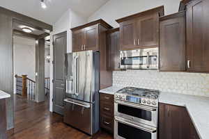 Kitchen featuring appliances with stainless steel finishes, dark brown cabinets, decorative backsplash, dark wood-style flooring, and vaulted ceiling