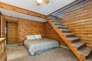 Carpeted bedroom with rustic walls, a textured ceiling, a baseboard radiator, ceiling fan, and beam ceiling
