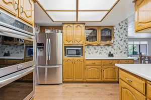 Kitchen with stainless steel appliances, light countertops, wallpapered walls, glass insert cabinets, and brown cabinets