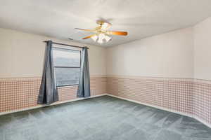 Empty room featuring wallpapered walls, a wainscoted wall, a textured ceiling, carpet flooring, and a ceiling fan