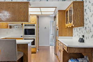 Kitchen with light countertops, glass insert cabinets, wallpapered walls, brown cabinetry, and stainless steel appliances