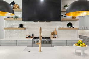 Kitchen with open shelves, range hood, gray cabinets, light stone counters, and backsplash