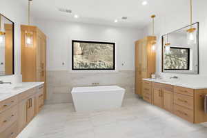 Full bathroom featuring tile walls, two vanities, light marble finish floors, a soaking tub, and wainscoting