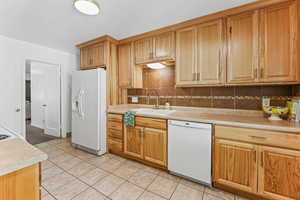 Kitchen featuring white appliances, decorative backsplash, light countertops, light tile patterned floors, and brown cabinetry