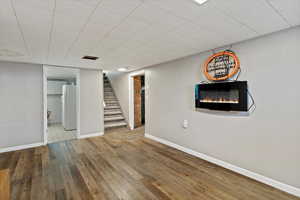 Finished basement featuring stairs, wood-type flooring, freestanding refrigerator, a glass covered fireplace, and a paneled ceiling