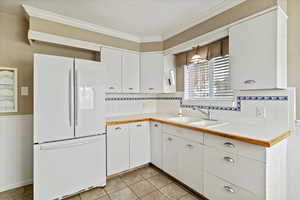 Kitchen with tile countertops, white cabinets, freestanding refrigerator, ornamental molding, and wainscoting