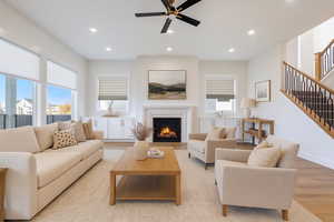 Living room with stairs, recessed lighting, a lit fireplace, wood finished floors, ceiling fan, natural light