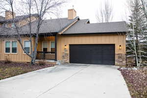 View of front of house featuring stone siding, a chimney, roof with shingles, concrete driveway, and covered porch