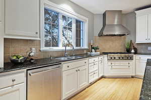 Kitchen with wall chimney range hood, dark stone counters, appliances with stainless steel finishes, and white cabinetry