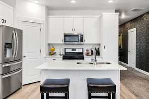 Kitchen with appliances with stainless steel finishes, white cabinets, black hardware, a kitchen breakfast bar, an island with sink, and light wood-style flooring