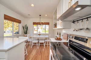 Kitchen with stainless steel range with electric cooktop, under cabinet range hood, white cabinetry, decorative light fixtures, and a kitchen bar