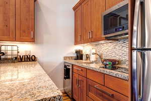 Kitchen featuring appliances with stainless steel finishes, brown cabinetry, backsplash, light stone countertops, and dark wood-style floors