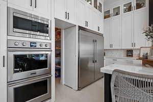 Kitchen featuring built in appliances, white cabinetry, light stone countertops, glass insert cabinets, and tasteful backsplash