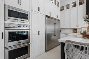 Kitchen featuring built in appliances, light stone countertops, white cabinets, backsplash, and glass insert cabinets