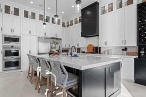Kitchen with light stone counters, an island with sink, white cabinetry, built in appliances, and decorative light fixtures