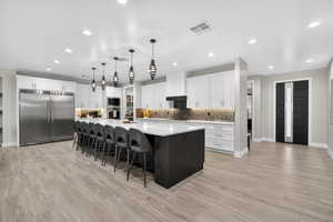 Kitchen featuring white cabinetry, a spacious island, built in appliances, a kitchen breakfast bar, and decorative light fixtures