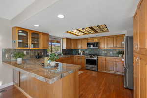 Kitchen with a peninsula, dark wood-style flooring, stainless steel appliances, brown cabinets, and tasteful backsplash