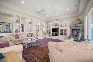 Living area featuring a ceiling fan, a raised ceiling, a chandelier, a stone fireplace, and built in shelves