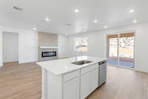Kitchen featuring light wood finished floors, a large fireplace, a center island with sink, stainless steel dishwasher, and white cabinetry